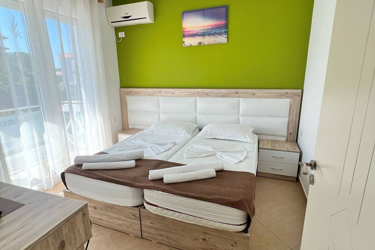 A bedroom with a bed, a bedside table, a nightstand, a wall-mounted air conditioner, a ceiling light, and a window with curtains.