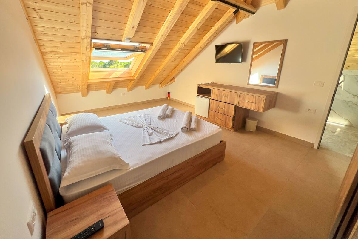 A bedroom with a wooden ceiling, a bed with white linens, a wooden nightstand, a small bedside table, a wooden dresser, a mirror, and a TV mounted on the wall.