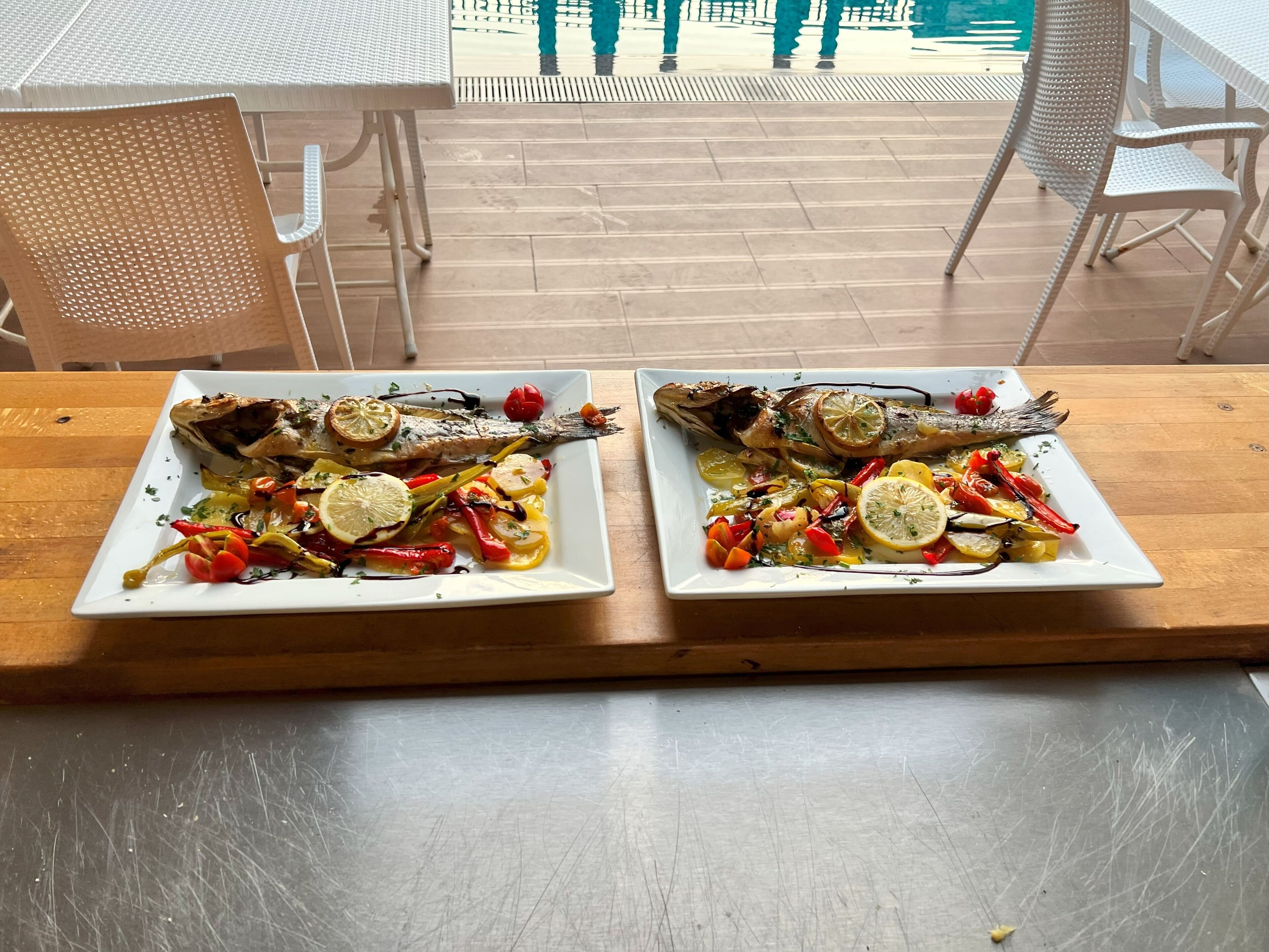 Two square white plates of roasted fish with lemon slices and vegetables on a wooden table.