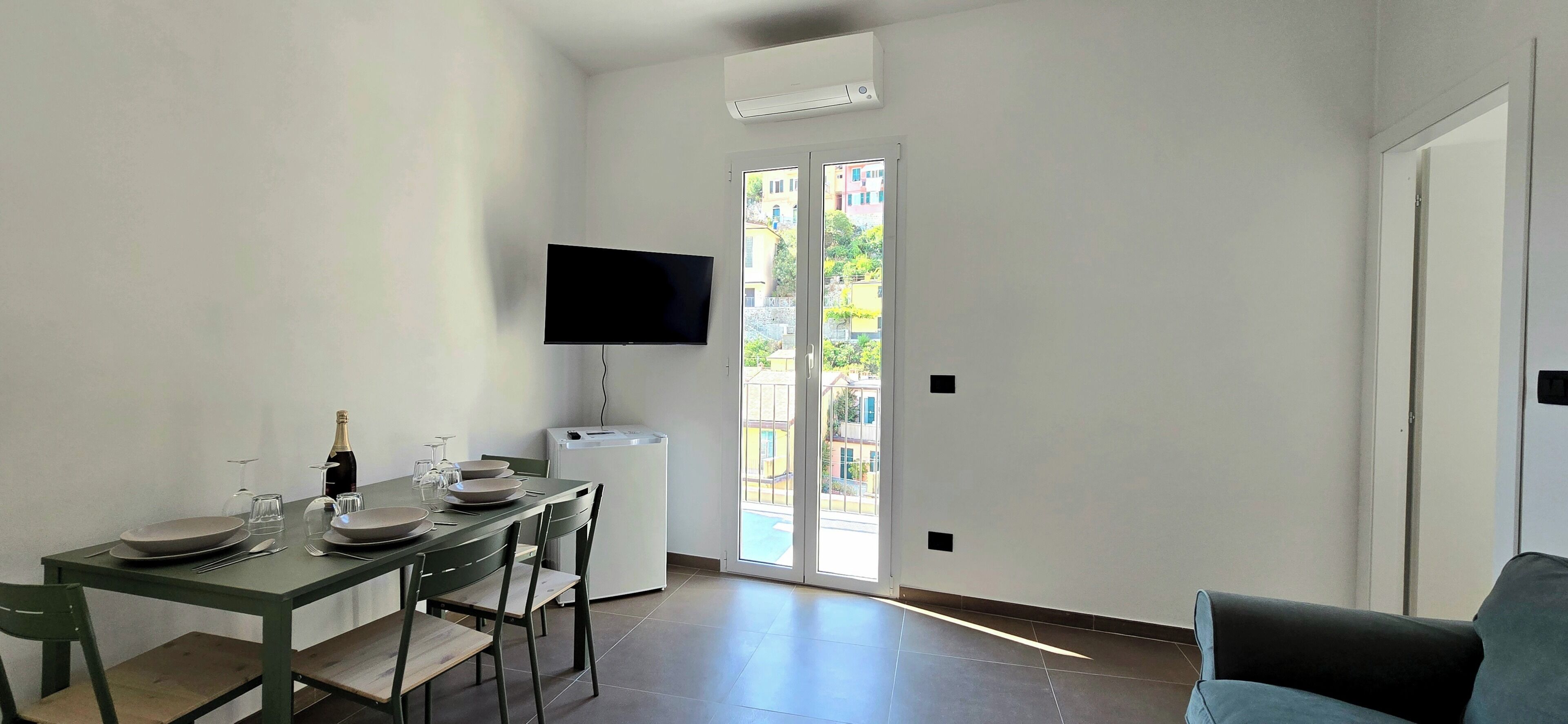 Un comedor con una mesa preparada para dos personas, un televisor de pantalla plana en la pared y un ventanal corredero que da a un balcón exterior.