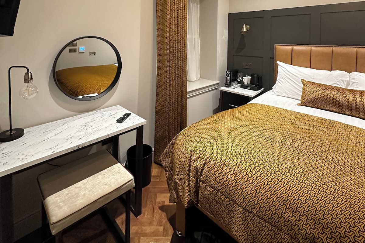A hotel room with a bed, a desk with a mirror, a round mirror, a bedside table, a lamp, and a ceiling light fixture.