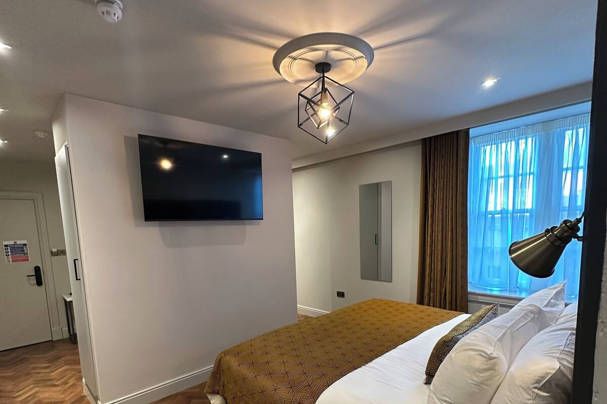 A modern hotel room with a large bed, a flat-screen TV mounted on the wall, a ceiling light fixture, and a window with curtains.