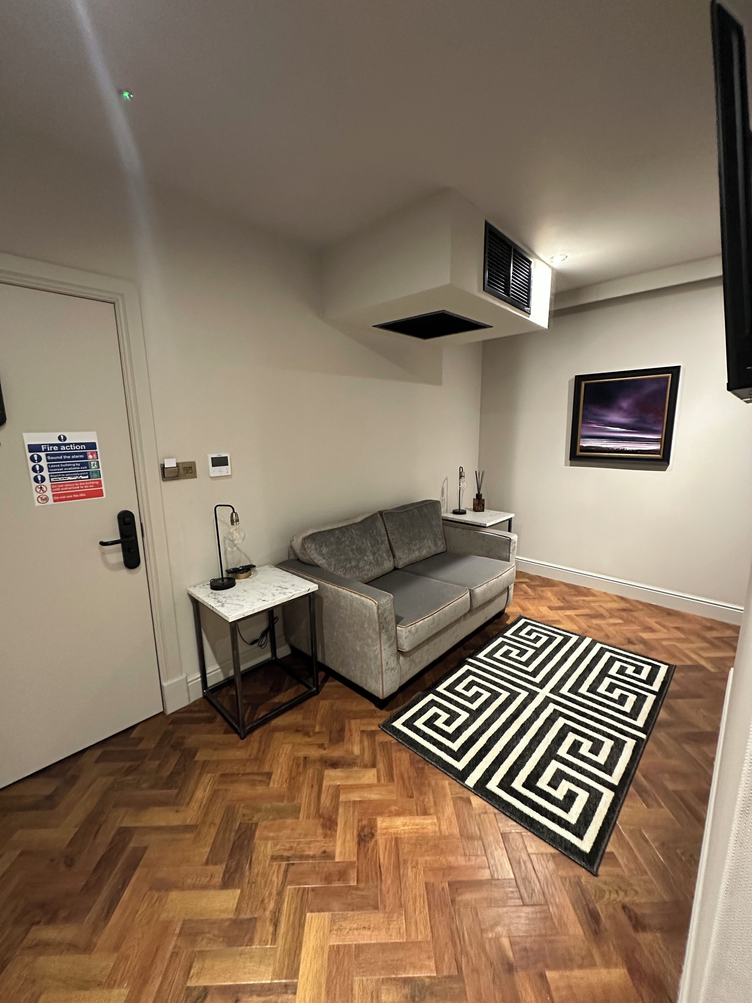 A room with a grey sofa, a small table with a lamp, a patterned rug, a framed picture on the wall, and a fire action sign on the door.