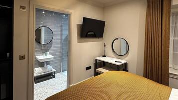 Una habitación de hotel moderna con una cama grande, una mesita de noche pequeña, un espejo, un televisor y un baño visible a través de una puerta abierta.