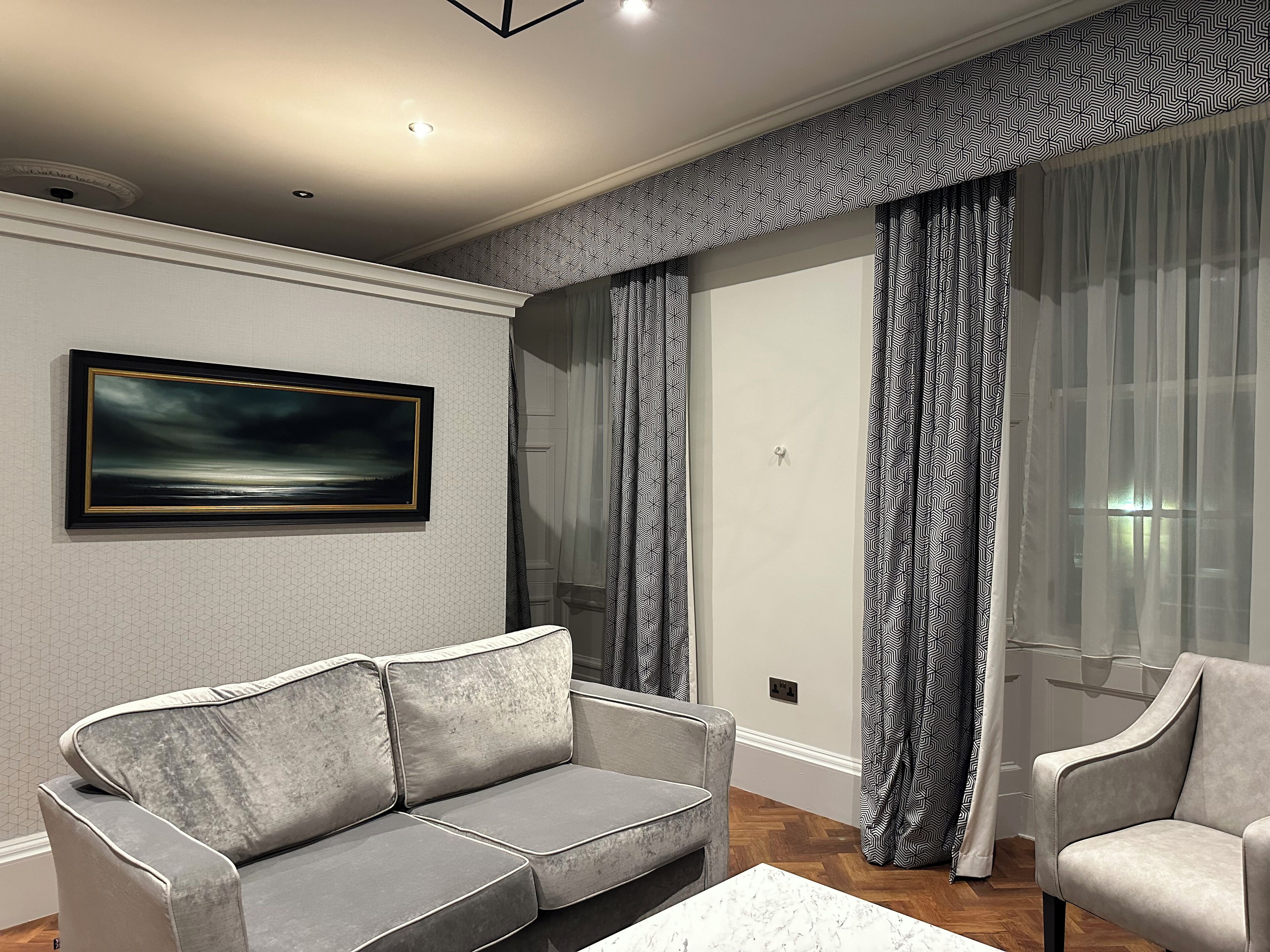 A modern living room with a grey sofa, a matching armchair, and a marble coffee table. A large abstract painting adorns the wall, and patterned curtains frame a window.