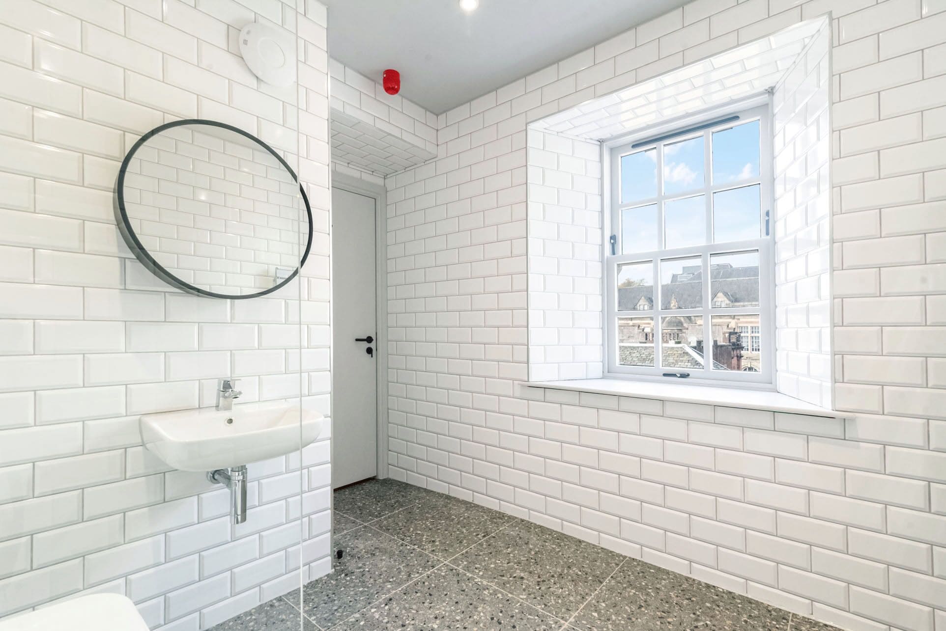 Un baño con paredes de ladrillo blanco, un espejo redondo, un lavabo y una ventana con vistas a edificios.