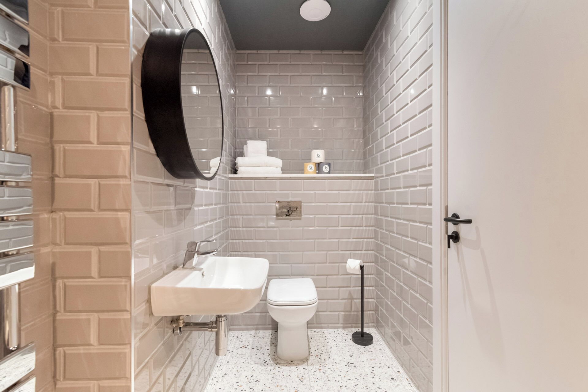 A bathroom with a white sink, toilet, and a mirror. There is a shelf with towels and toilet paper. The walls are tiled with a brick pattern.