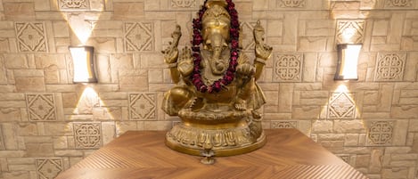 A statue of a deity on a wooden table with a patterned stone wall in the background.