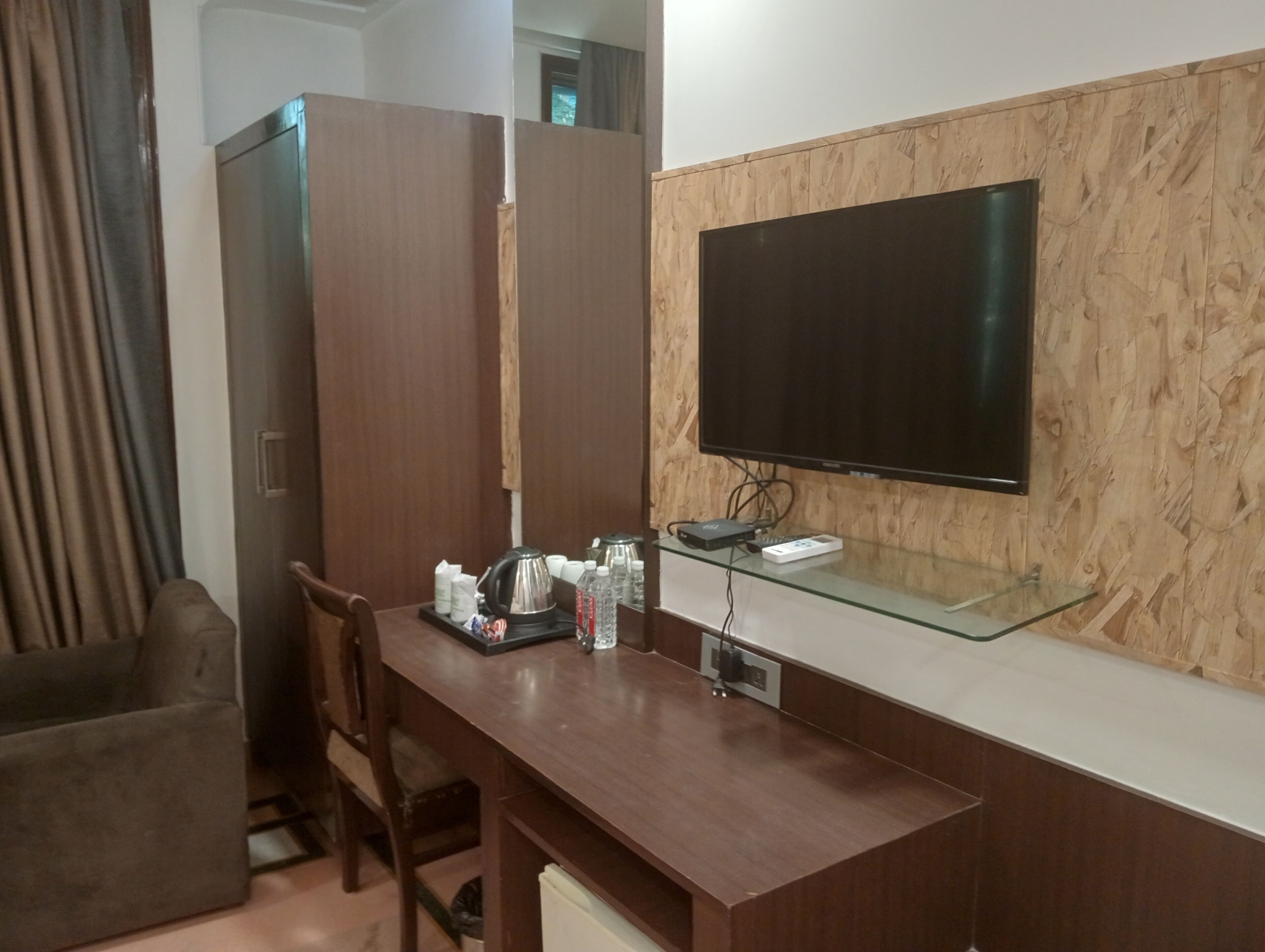 Ein Hotelzimmer mit einem Holztisch, einem Stuhl, einem an einer Holzplatte montierten Fernseher und einem Glastischchen mit Steckleiste und Fernbedienung.