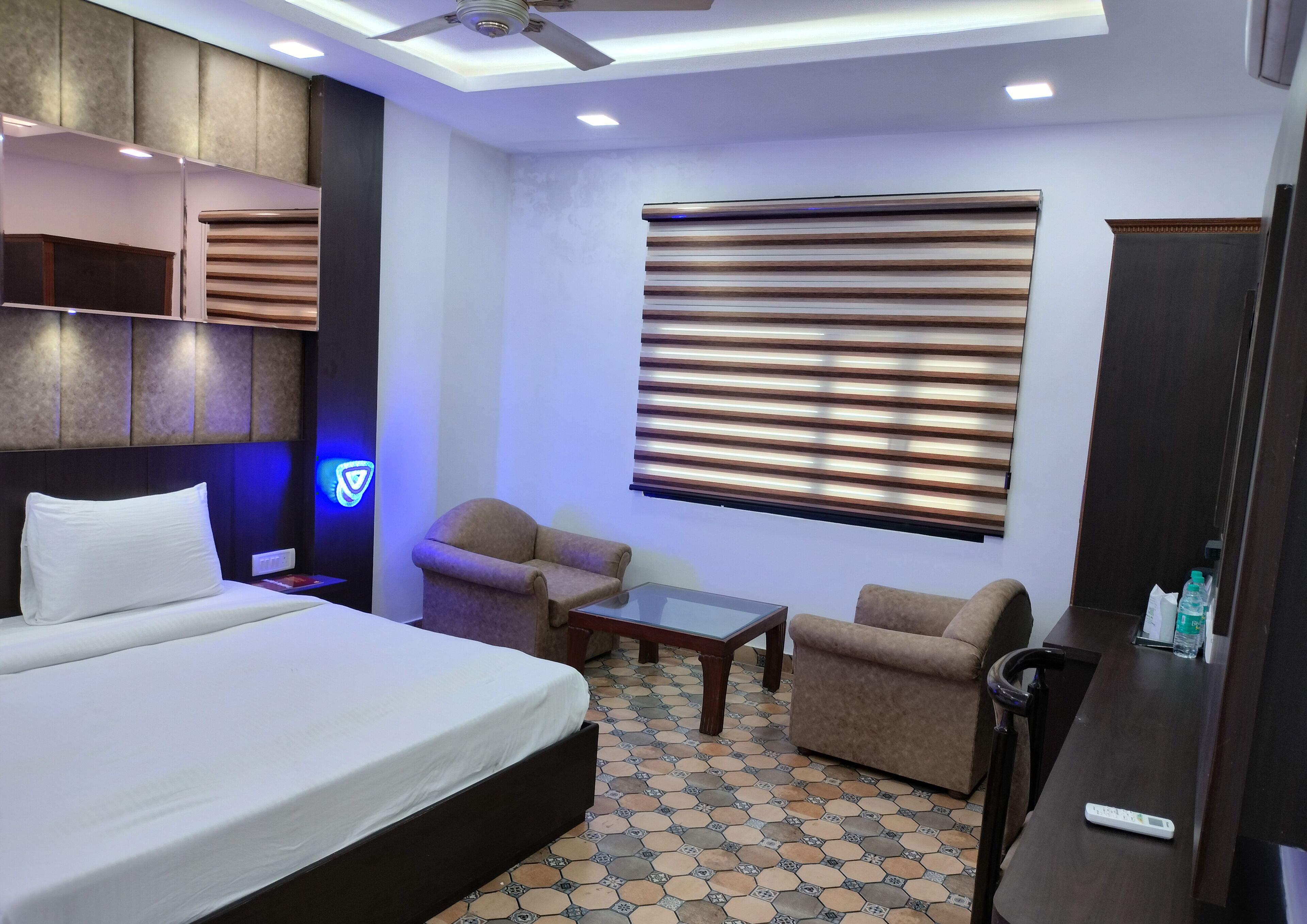 Habitación de hotel con una cama grande, dos sillones, una mesita, un ventilador de techo y una ventana con persianas.