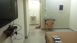 A hotel room with a bed, a desk with a chair, a TV mounted on the wall, and a bathroom visible through an open door. - Orchid Residency Medavakkam (Chennai)
