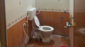 A small bathroom with a toilet, a shower, and a red bucket. - Orchid Residency Medavakkam (Chennai)