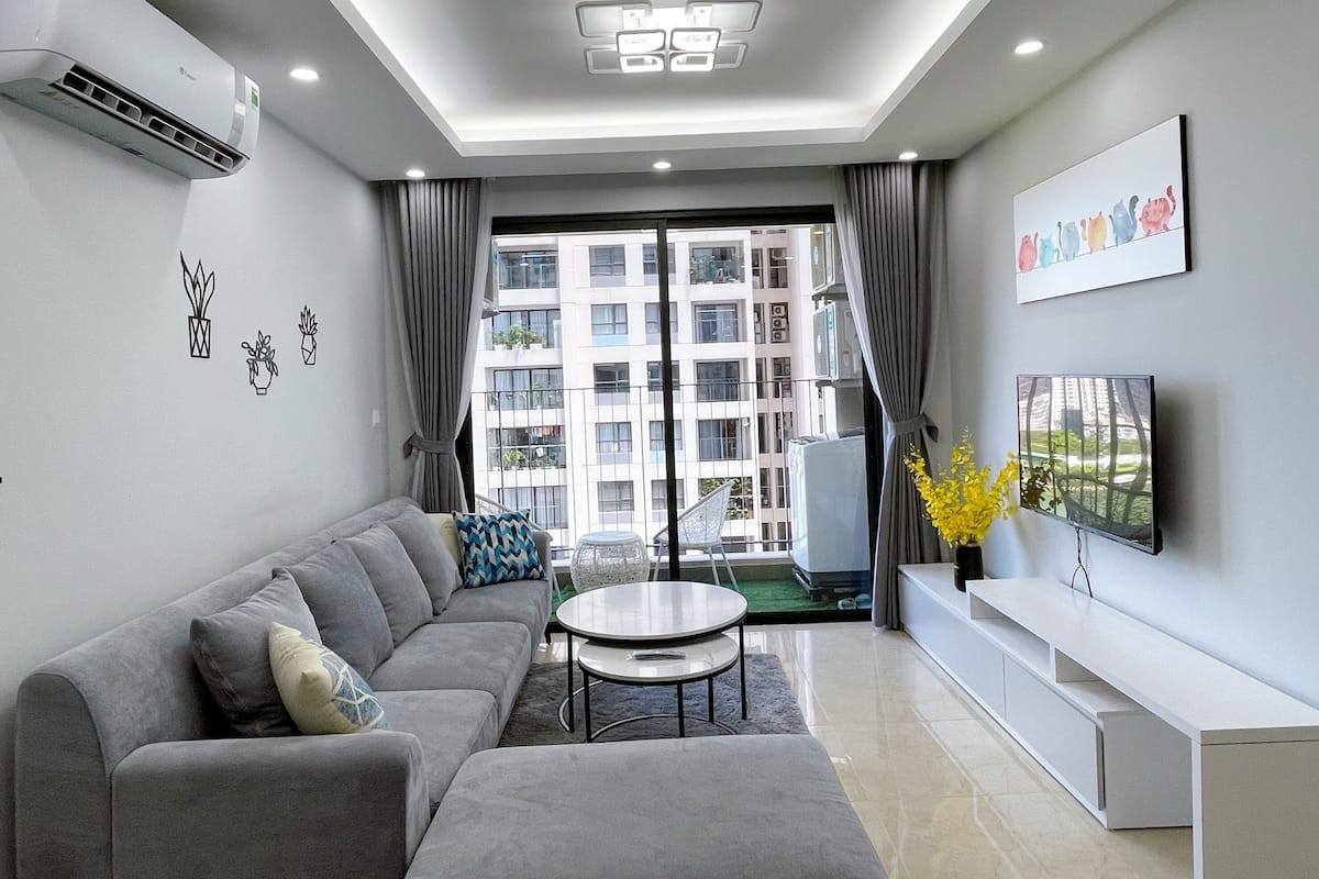 A modern living room with a grey sofa, a coffee table, a TV on a white stand, and a balcony with a view of buildings.