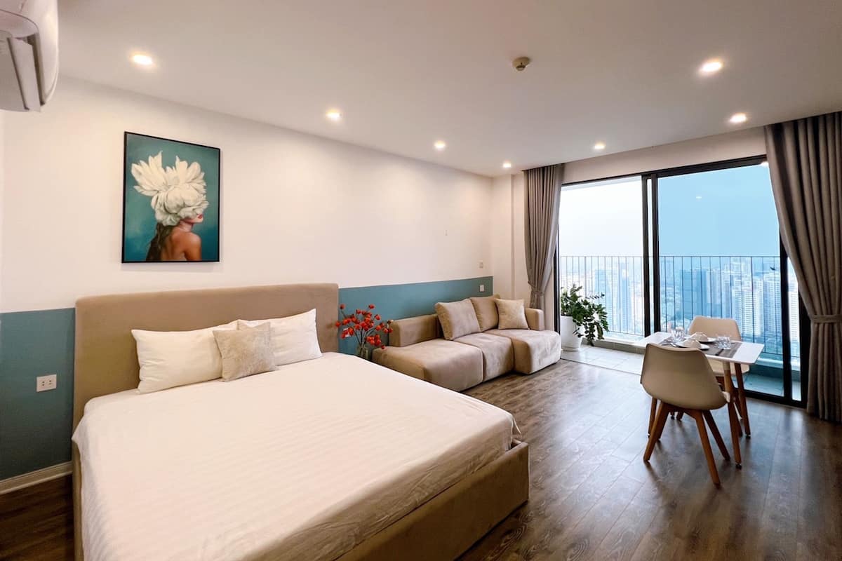 A modern hotel room with a large bed, a sofa, a dining table, and a view of the city through large windows.