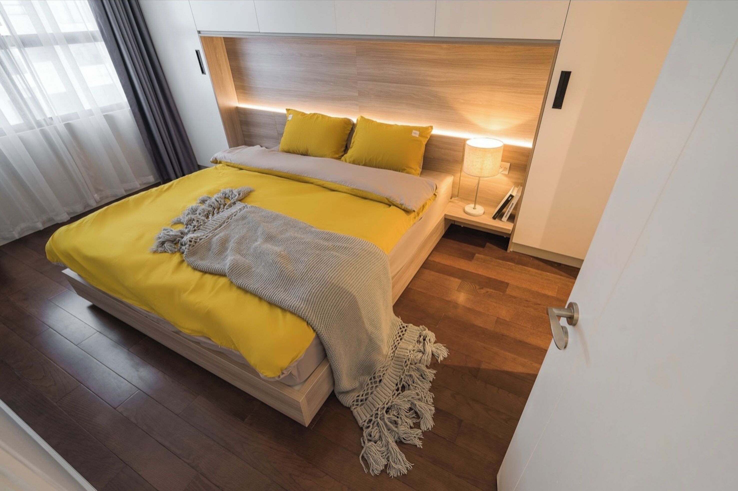 A modern bedroom with a wooden floor, a bed with yellow bedding, a bedside table with a lamp, and a wooden headboard.