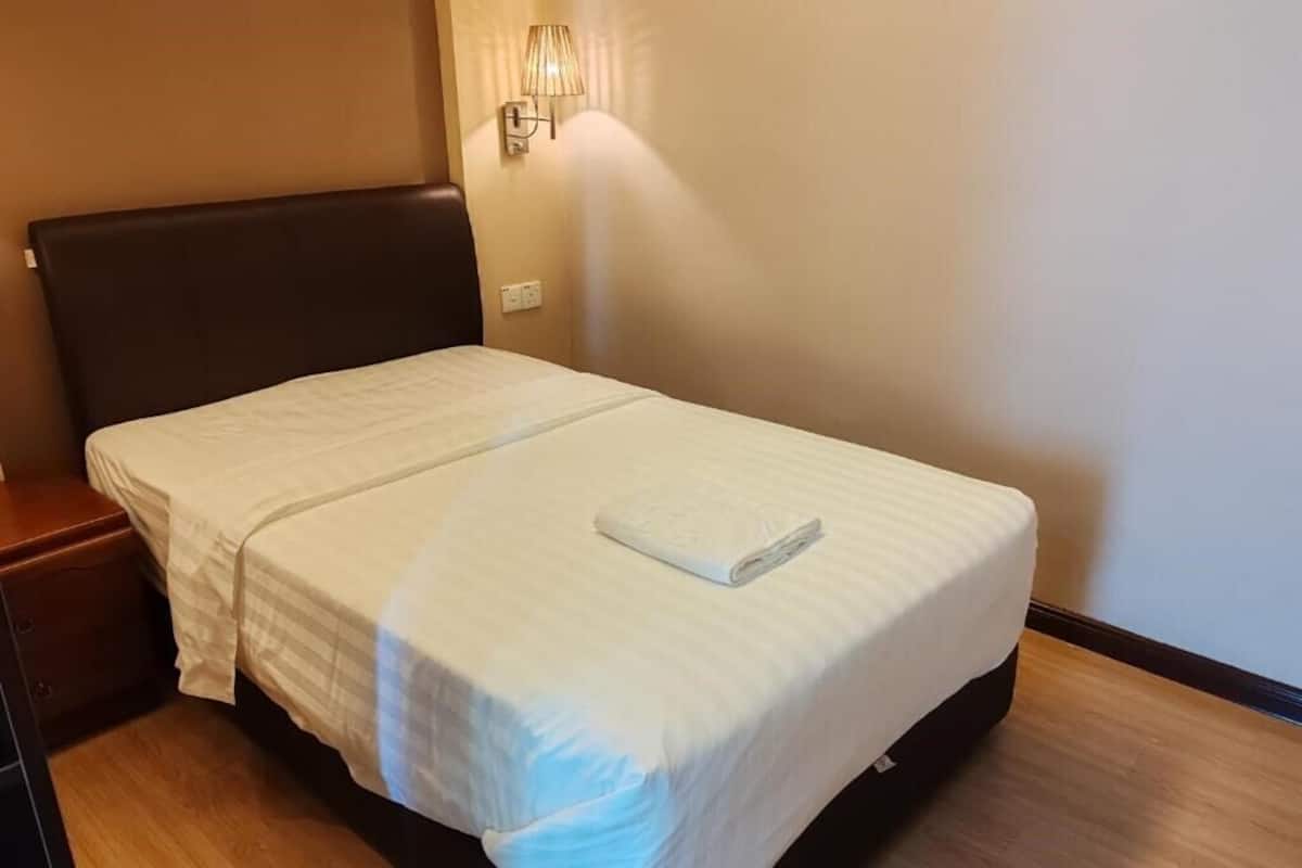 A neatly made bed with a white bedspread and a folded towel, a wooden nightstand, and a wall-mounted lamp.