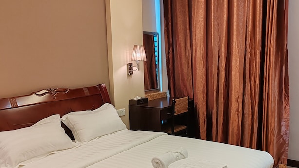 Hotellihuone, jossa on suuri sänky, puinen pääty, yöpöytä, lamppu ja ikkunasta avautuva näkymä ulos, ikkunassa on verhot.