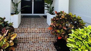 Un ingresso coperto con un vialetto piastrellato che conduce a una porta nera, circondato da piante in vaso e da un muro bianco.