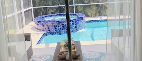 En utsikt från ett rum med en glasdörr som vetter mot ett poolområde, där det finns en bubbelpool och solstolar.