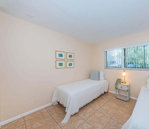 Un lit simple avec une couvre-lit blanche, dans une pièce aux murs de couleur beige, une fenêtre avec des stores et une petite table de chevet avec une lampe.