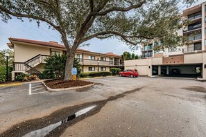 Un edificio a più piani con un'area di parcheggio, un'auto rossa parcheggiata nel parcheggio e un albero sullo sfondo.