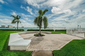Area esterna pavimentata con palme, una panchina a forma di curva e vista su edifici attraverso uno specchio d'acqua.