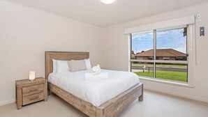 A bedroom with a wooden bed, white bedding, a nightstand, and a large window with a view of a residential area.