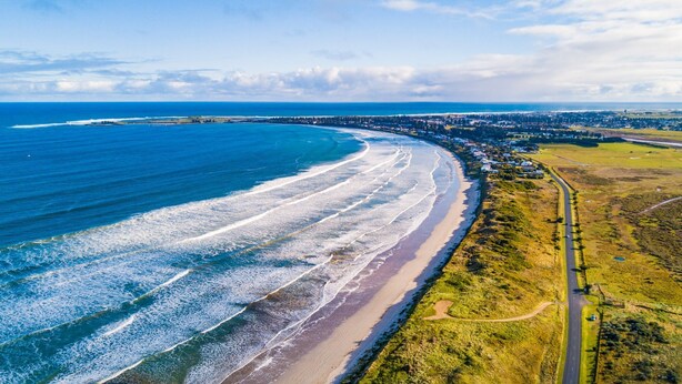 An aerial view of a coastal area with a beach, a road, and a town in the background.