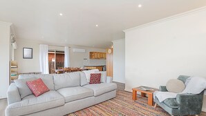 A modern living room with a grey sofa, a small wooden table, and a green armchair. There is a kitchen in the background with wooden cabinets and a brick wall.