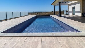 Una piscina rectangular con agua azul cristalina, rodeada por un solárium con baldosas y una barandilla de vidrio.