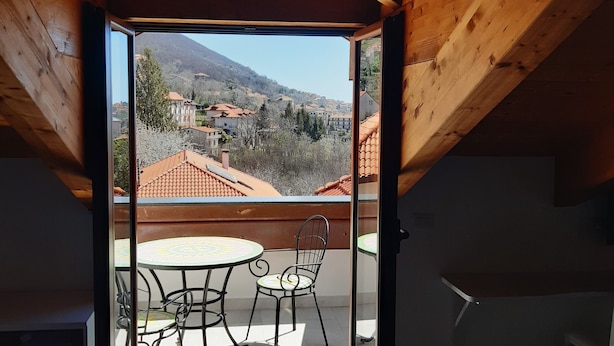 Um quarto de sótão com teto de madeira, porta de vidro que dá para uma varanda com mesa e cadeiras, e vista para uma aldeia com casas e colinas.