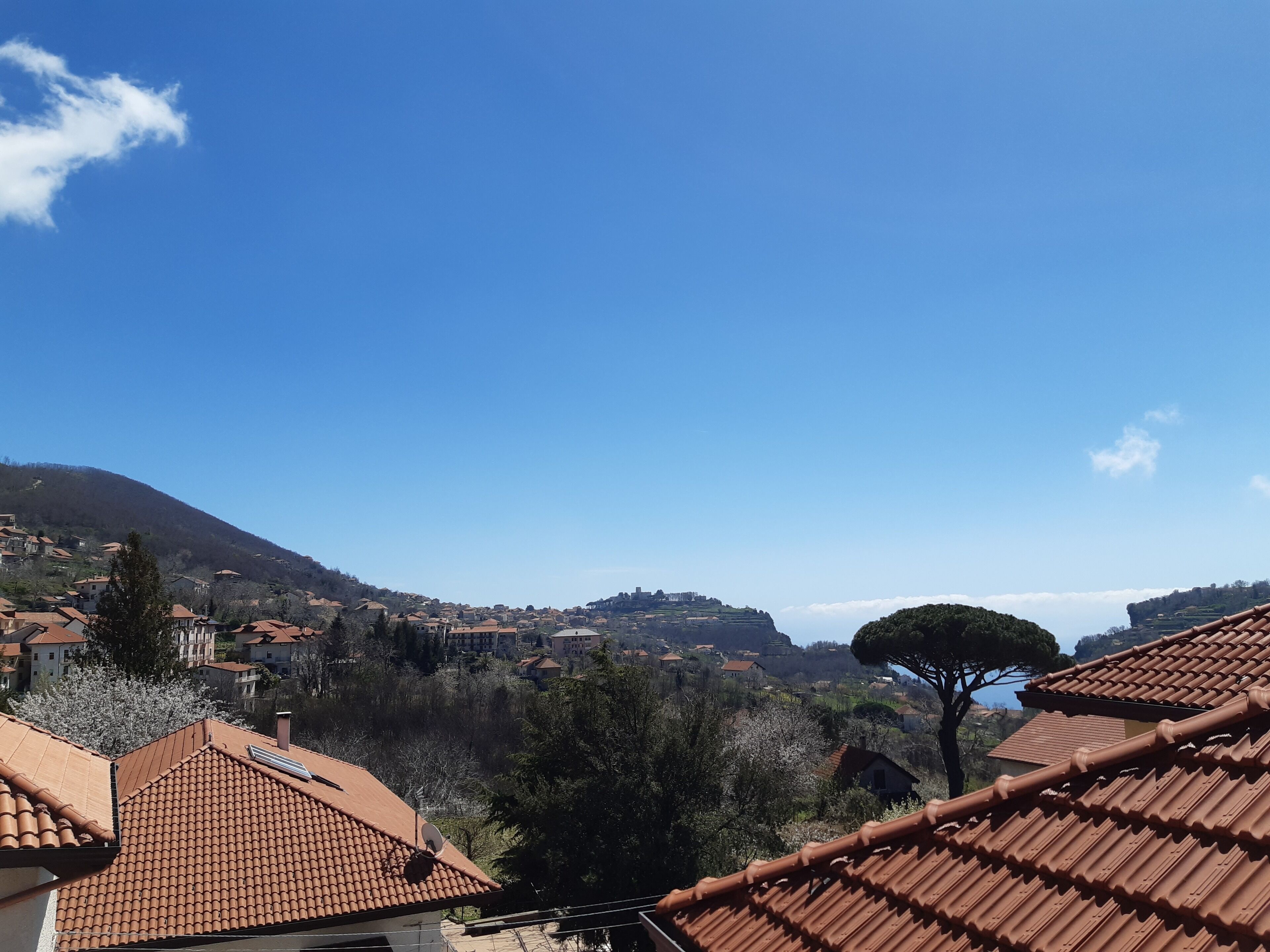 Un village perché sur une colline, avec des bâtiments aux toits de tuiles rouges, un arbre imposant et un ciel d’un bleu éclatant.