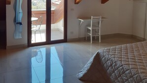 Quarto com teto inclinado, uma cama, uma escrivaninha e vista através de uma porta de vidro.