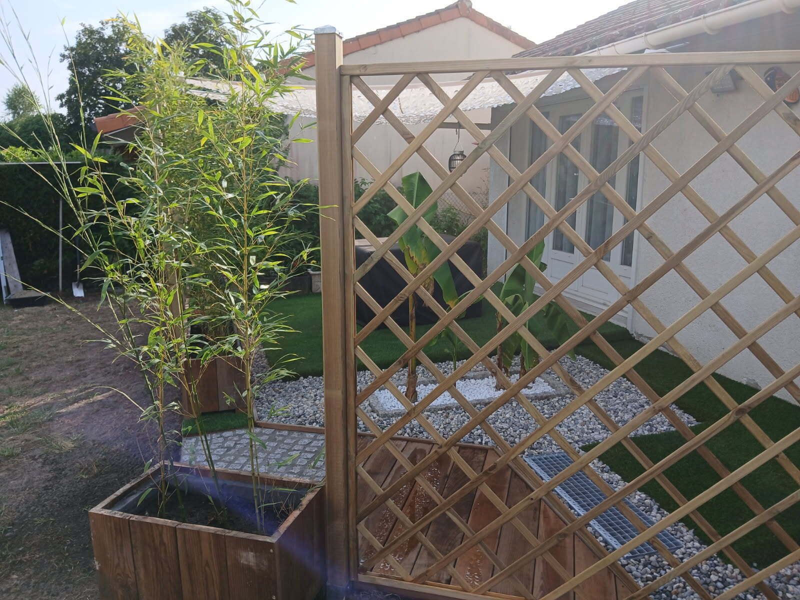 Een houten hek met een lattenwerk, een pot met bamboe en een kleine tuin met grind en gras.