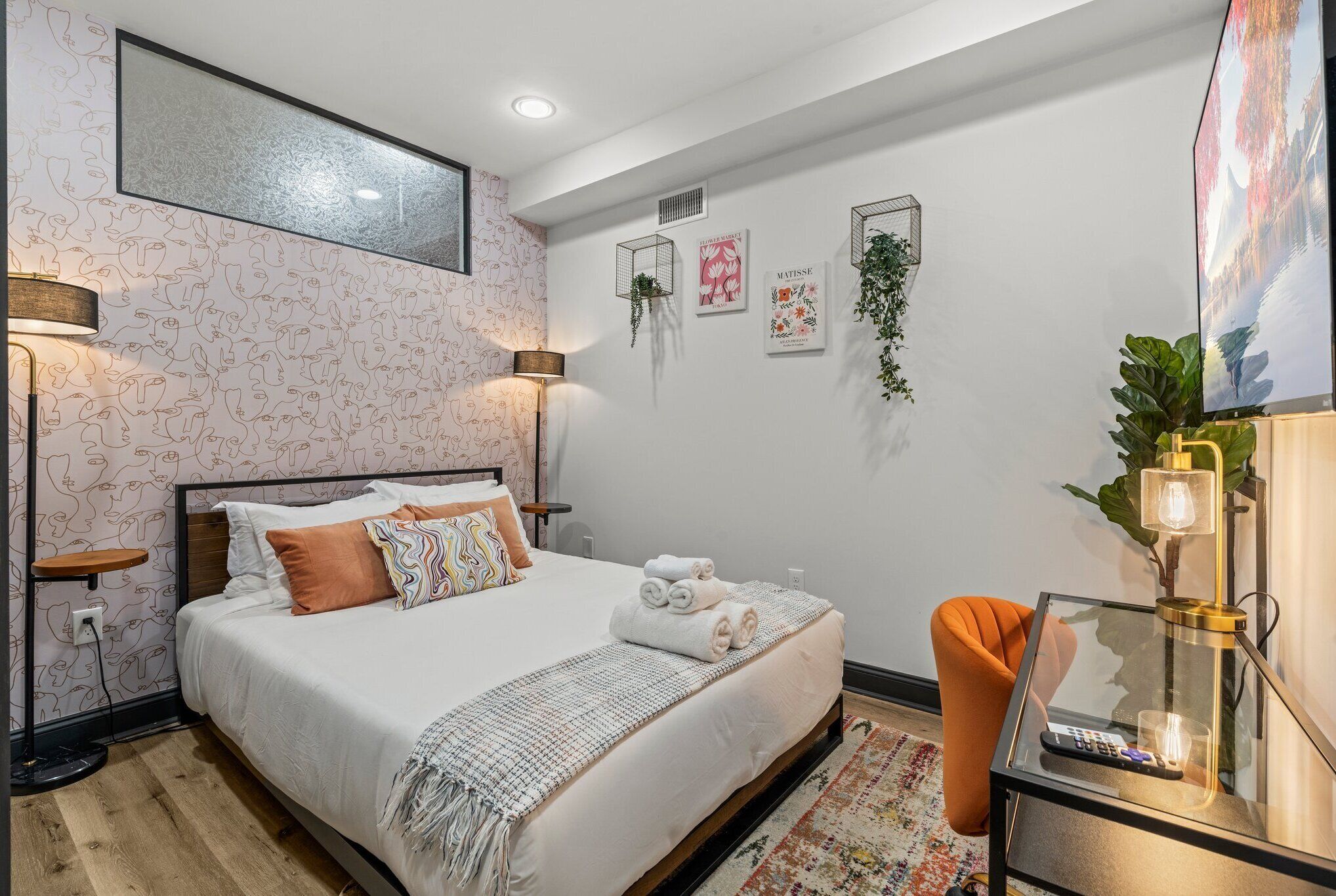 A neatly made bed with white linens and a decorative throw, a bedside table with a lamp, and a small orange chair.