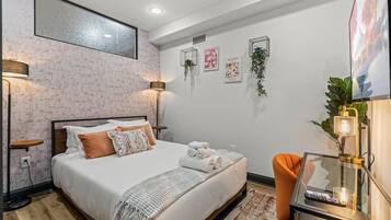 A neatly made bed with white linens and a decorative throw, a bedside table with a lamp, and a small orange chair.