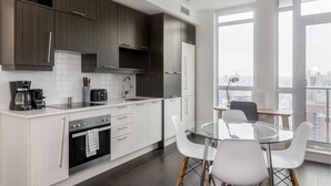 Una cocina moderna con armarios blancos, horno empotrado y comedor con mesa de vidrio y sillas blancas.