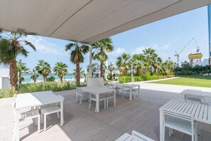 Een terras buiten met witte tafels en stoelen, palmbomen en uitzicht op de oceaan en bouwkranen.