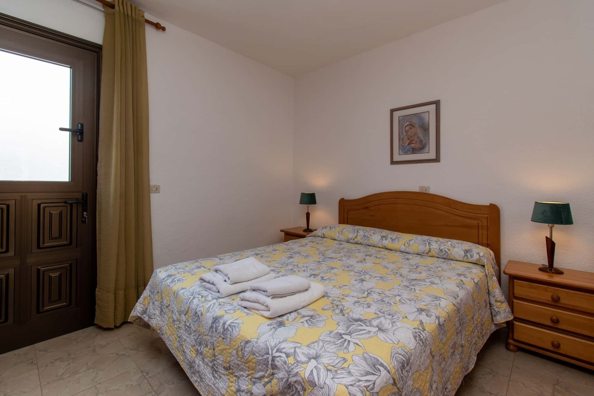 Una cama bien hecha con ropa de cama floral, cabecera de madera, dos mesitas de noche con lámparas, una puerta con cortina y un cuadro en la pared.