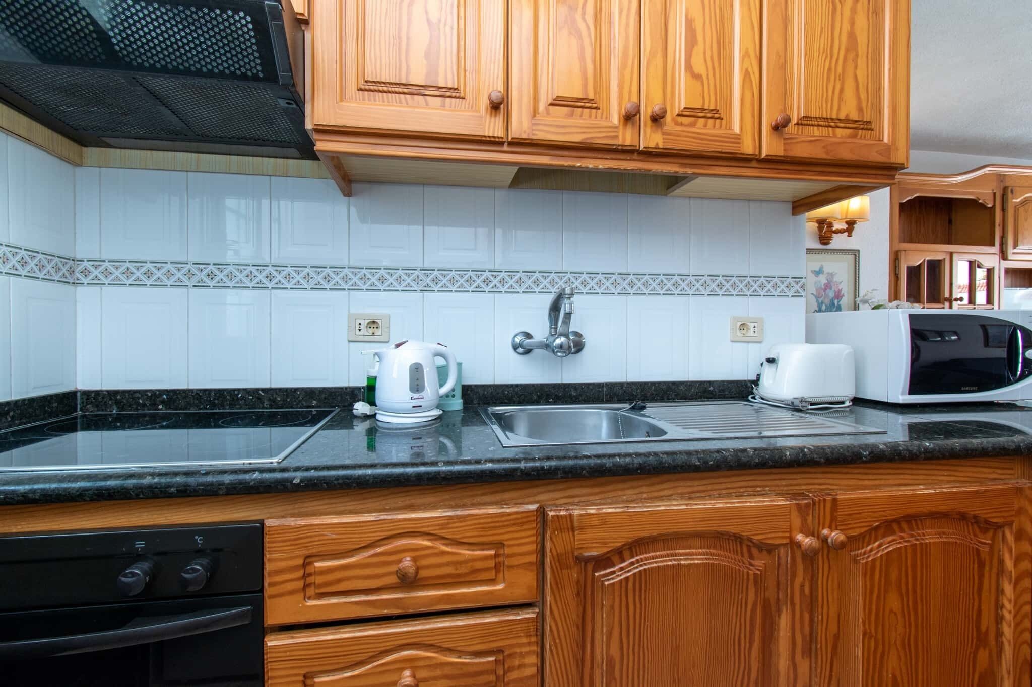 A kitchen with wooden cabinets, a black countertop, a stainless steel sink, a kettle, a toaster, and a microwave.
