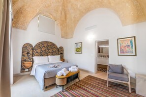 Une chambre à coucher comprenant un lit, une table de chevet, une chaise, une porte donnant sur une salle de bain et un mur orné d’un tableau.