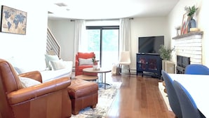 A living room with a sofa, ottoman, armchair, and a small table. There is a fireplace, a TV, and a staircase leading upstairs.