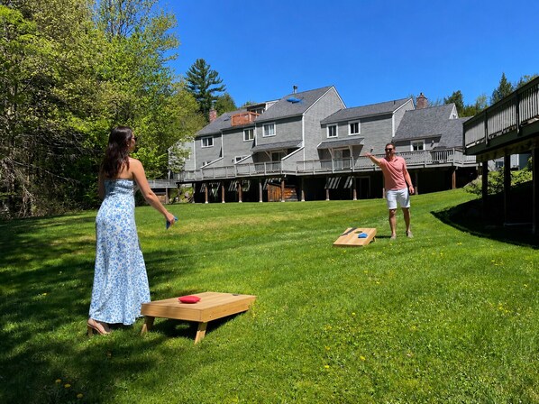 A grassy backyard with a wooden table and a red cushion, a house with a balcony in the background.