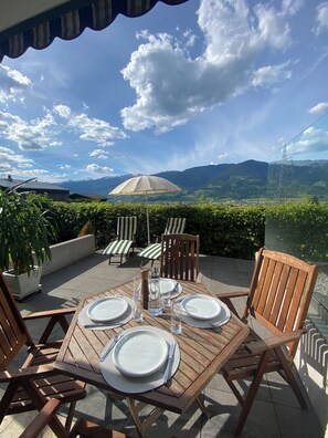 Un espace de restauration en plein air avec une table dressée pour quatre personnes, entourée de chaises en bois, offrant une vue sur un paysage montagneux.