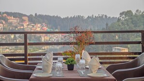 Mesa lista con cristalería transparente, platos y servilletas, con un centro de mesa de planta en maceta, con vistas a un paisaje de un pueblo y colinas boscosas.