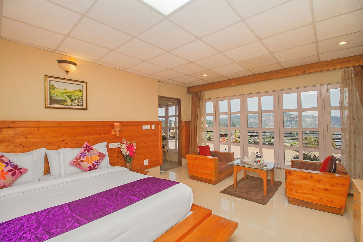 Habitación de hotel con una cama grande, cabecera de madera, un escritorio, un sofá y ventanas amplias con vistas.