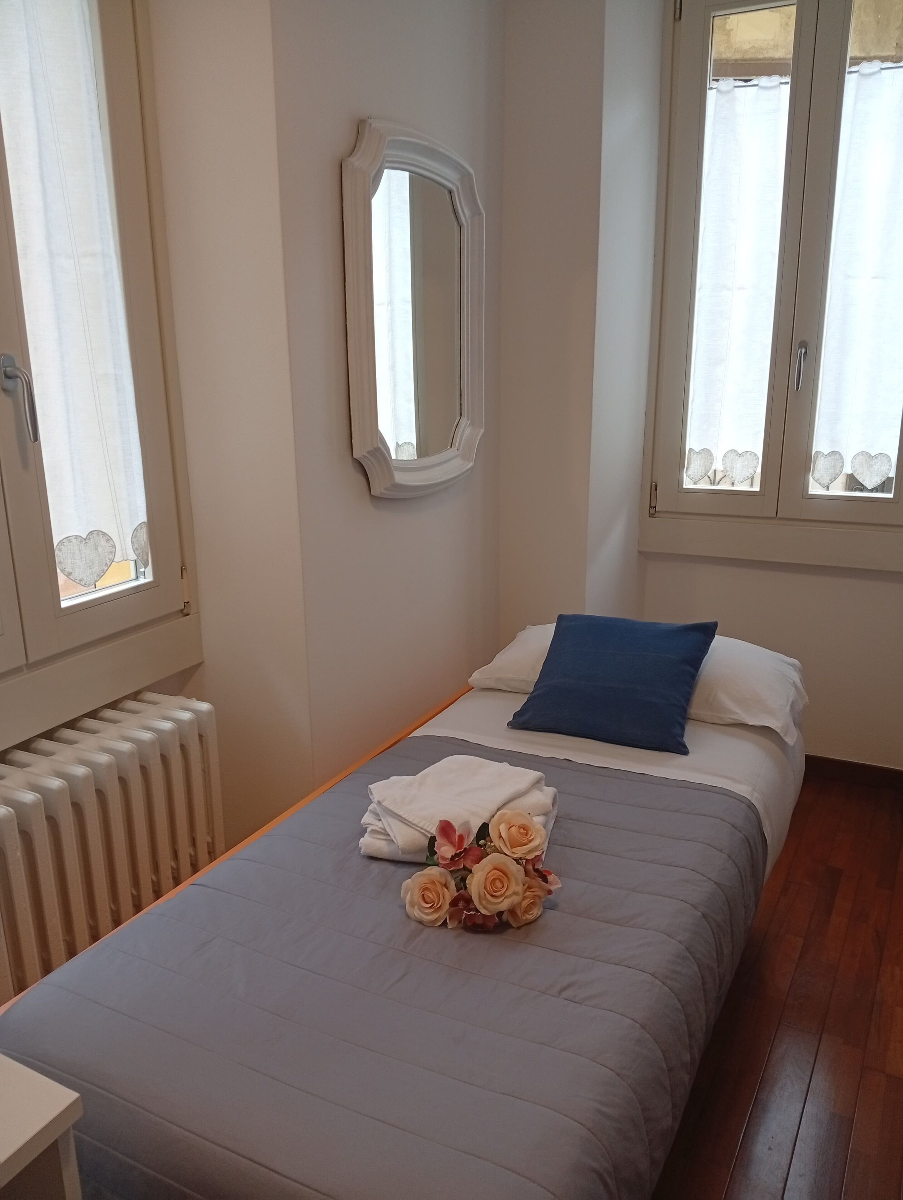 Ein ordentlich bezogenes Bett mit blauem Kopfkissen und einem Blumenstrauß, ein Heizkörper und ein Spiegel an der Wand.