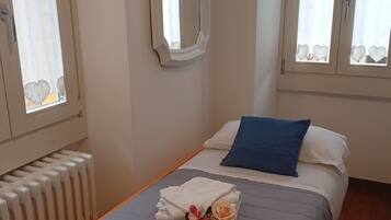 Ein ordentlich bezogenes Bett mit blauem Kopfkissen und einem Blumenstrauß, ein Heizkörper und ein Spiegel an der Wand.