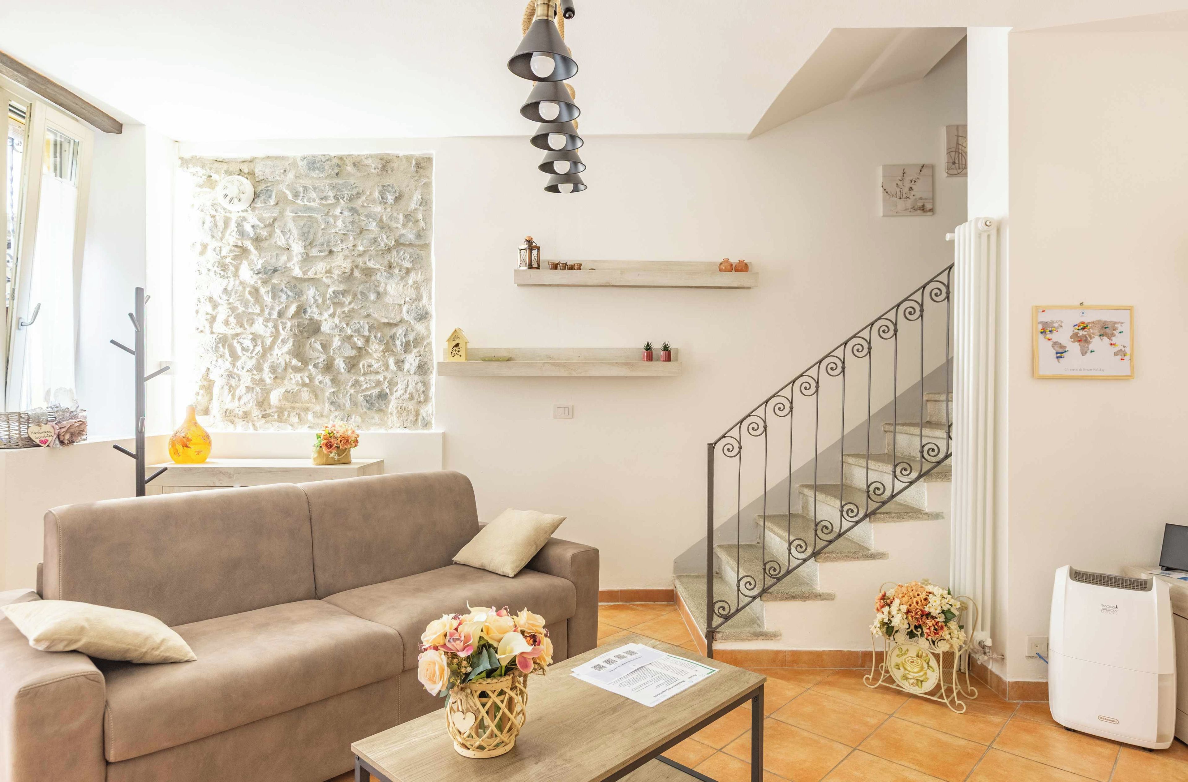 Ein Wohnzimmer mit einer Couch, einem Couchtisch mit Blumenvase, einer Treppe mit Geländer und einem an der Wand befestigten Regal mit Dekoration.