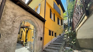 A narrow alleyway with a stone archway leading to a yellow building with a metal gate and a staircase. - Dream Holiday  (Argegno)